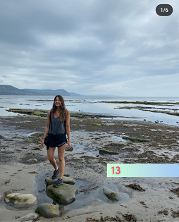 A white female with long brown hair smiles at the camera standing on a large rock at the waters edge